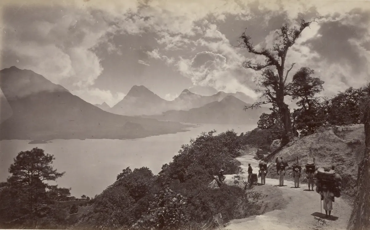 El Lago de Atitlán desde un camino en la ladera, con volcanes al otro lado del agua y una fila de cargadores en el camino, 1875.