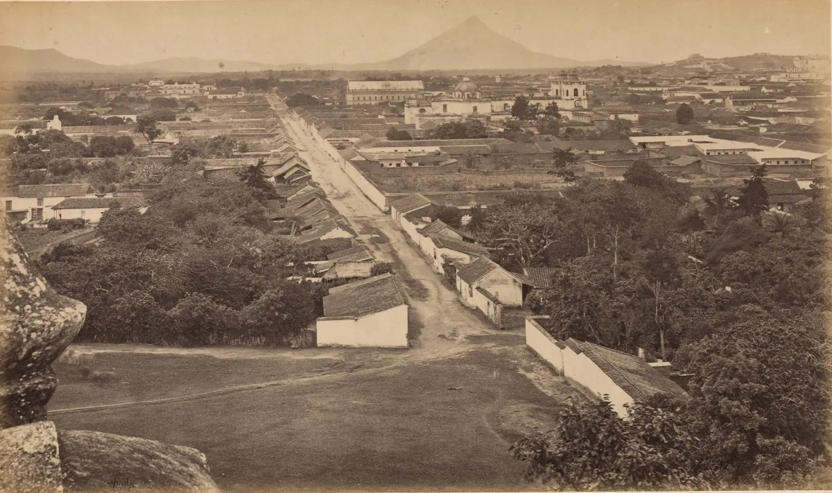 Vista desde una loma en 1875: una calle recta baja entre casas de teja hasta el cono del Volcán de Agua en el horizonte.
