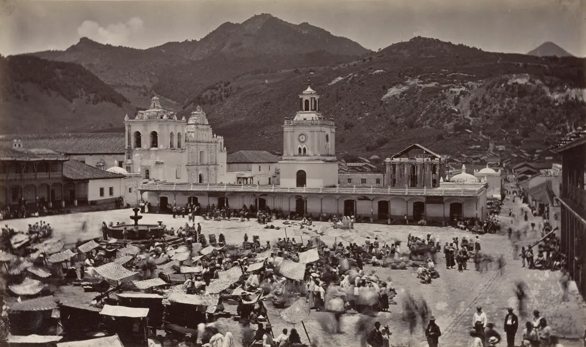 El parque central de Quetzaltenango en día de mercado, 1875: toldos de petate, mucha gente, la catedral y una torre con reloj, cerros detrás.