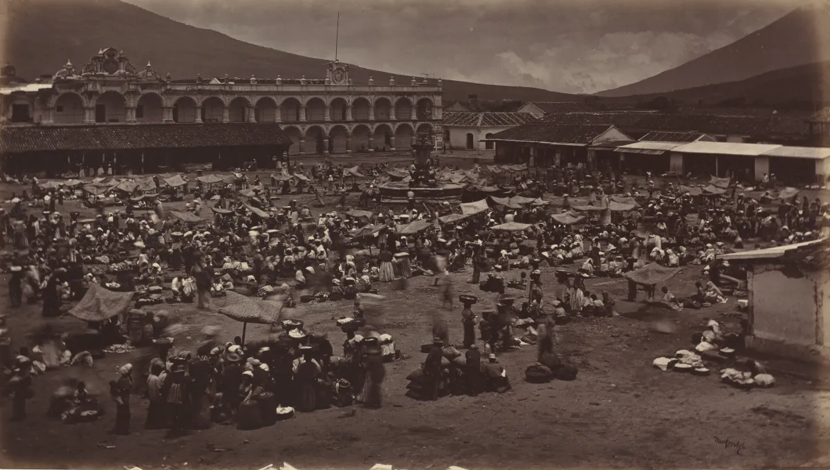 La Plaza Mayor de Antigua Guatemala llena de gente en día de mercado, 1877, con el Palacio de los Capitanes Generales al fondo y un volcán detrás.