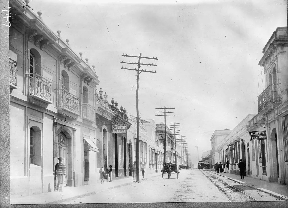 Una calle recta de la Ciudad de Guatemala en 1911, con balcones de hierro, postes de telégrafo, rieles de tranvía y un coche de caballos a lo lejos.